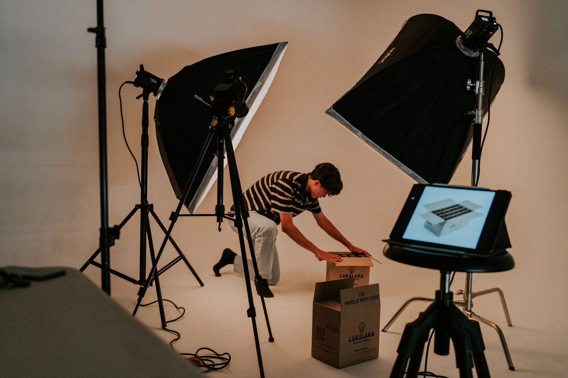 Un uomo prepara dei pacchi in uno studio con softbox, cavalletti e un tablet appoggiato su uno sgabello.