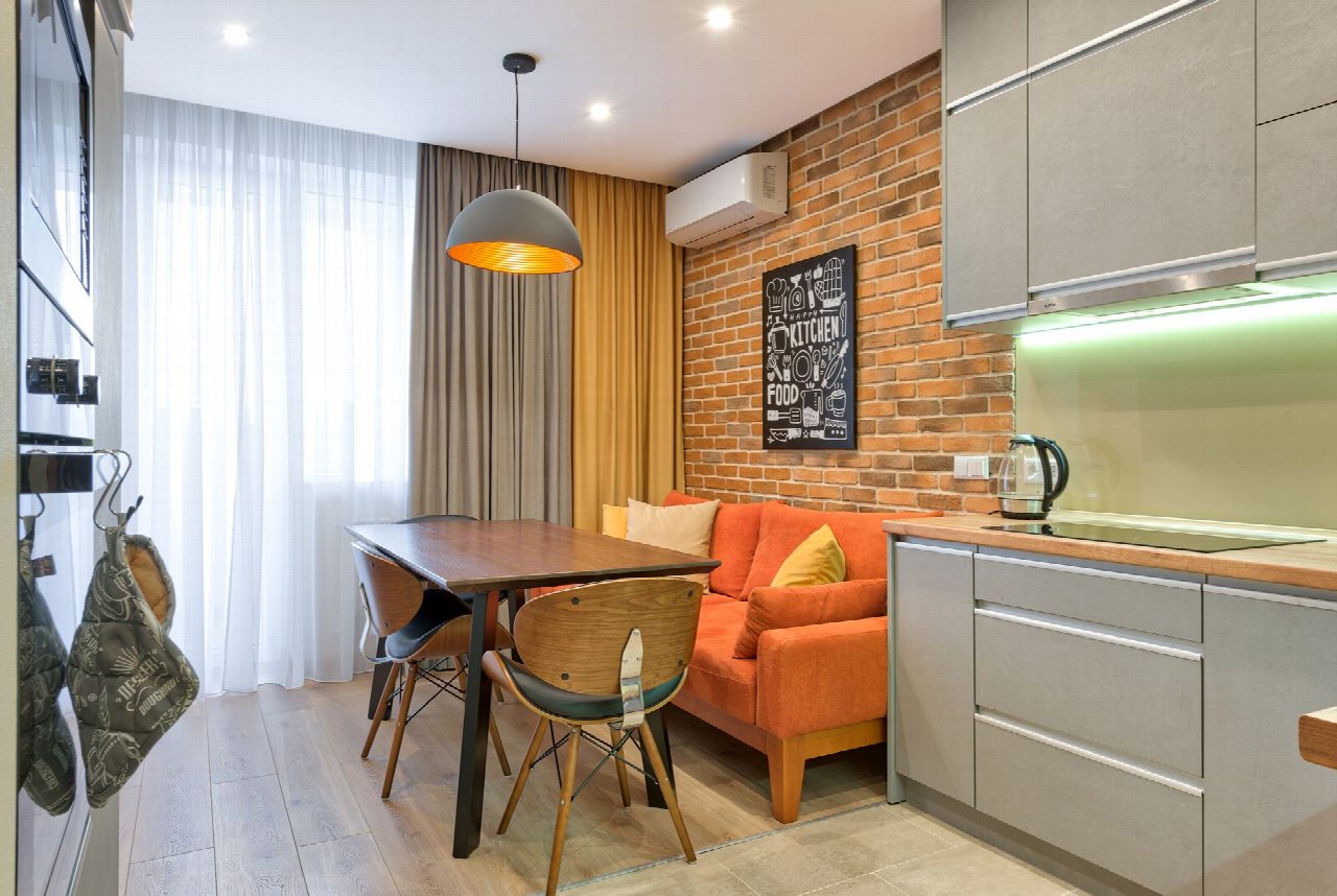 Cocina comedor con pared de ladrillo visto, sofá naranja, mesa de madera con sillas y encimera gris iluminada bajo los armarios.