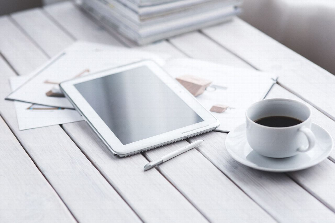 Tableta blanca con lápiz táctil, una taza de café y varias hojas sobre una mesa de madera clara.