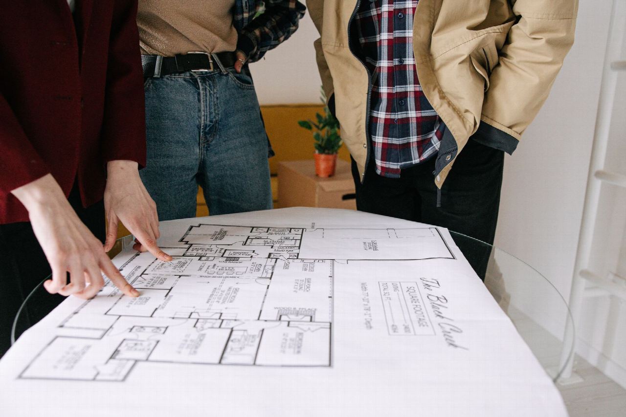 Varias personas de pie alrededor de una mesa de cristal, señalando el plano de una vivienda extendido sobre ella.